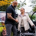 Ein Pfleger begrüßt eine Seniorin, die mithilfe eines Rollators aus dem Fahrdienst aussteigt.