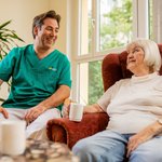Ein Pfleger sitzt neben einer Bewohnerin. Die beiden lächeln sich an.