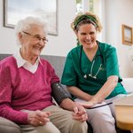 Lachende Seniorin sitzt auf einer Couch, eine Pflegerin misst ihren Blutdruck.